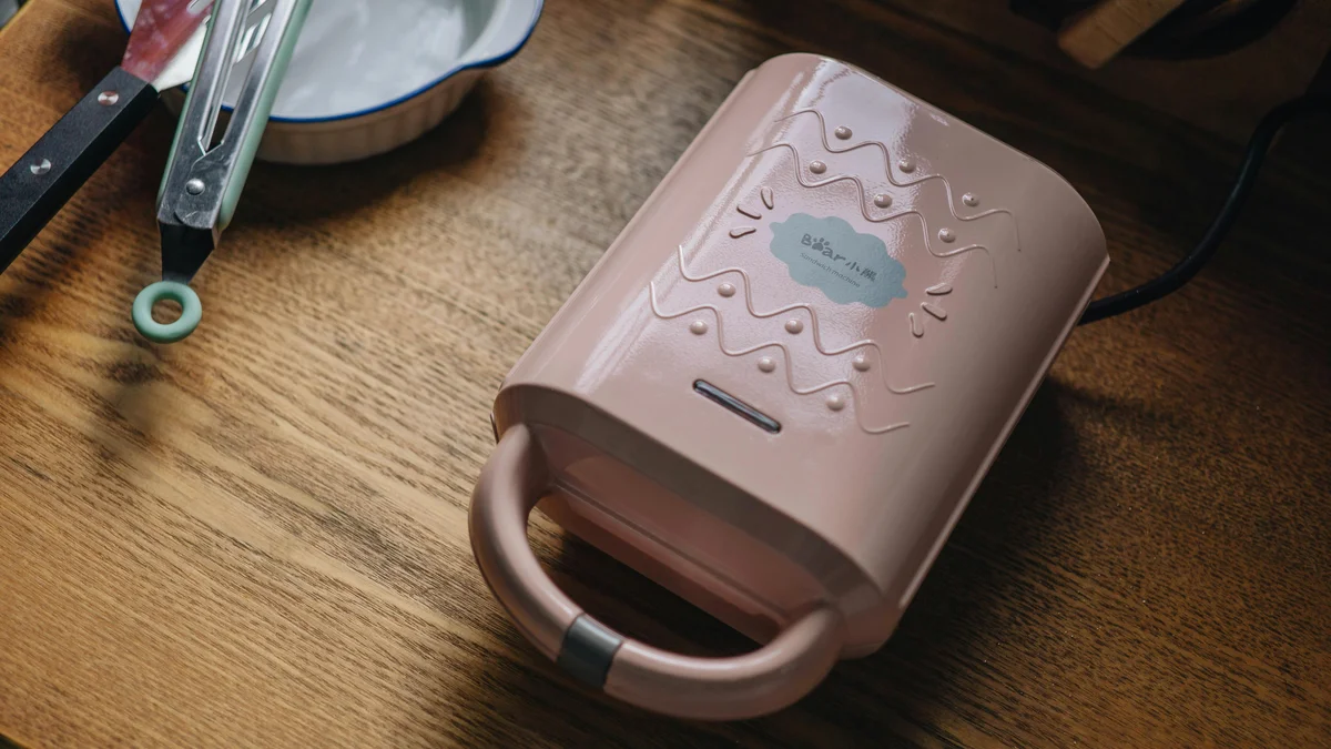 Pink sandwich maker on wooden table with kitchen utensils, perfect for cozy and modern kitchen setting.