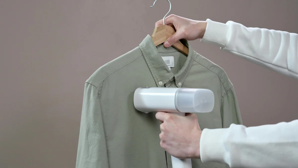 Mãos passando a vapor uma camisa verde com um vaporizador portátil contra um fundo cinza.