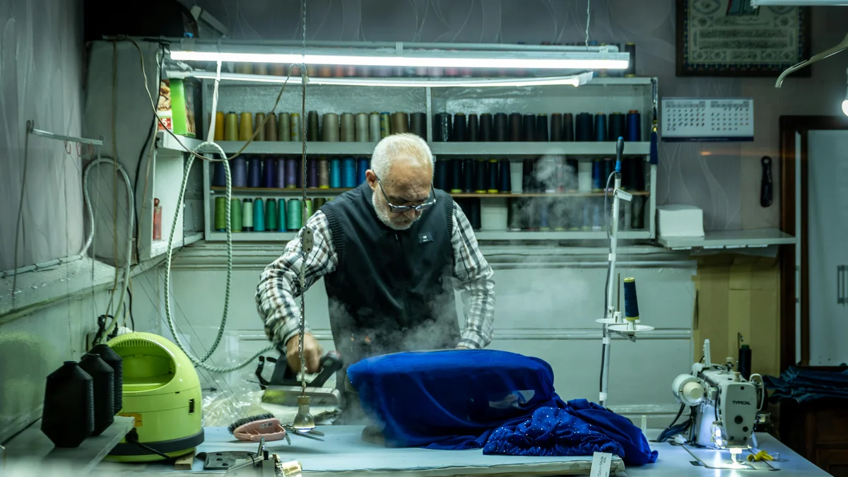 Alfaiate idoso passando tecido azul em oficina de costura tradicional cercado por ferramentas.