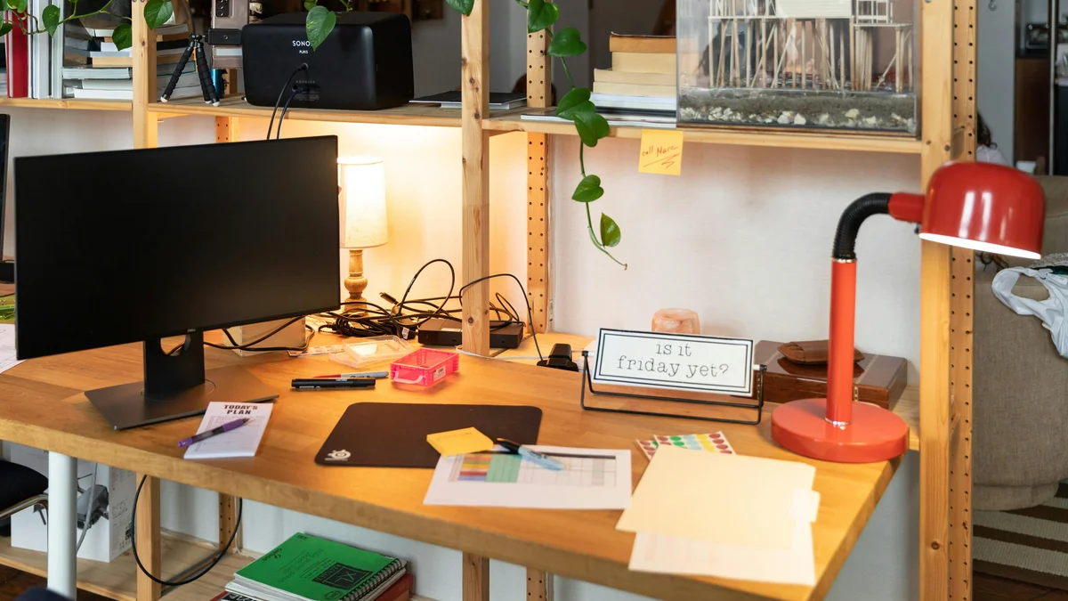 Espaço de trabalho aconchegante com escrivaninha de madeira, monitor, luminária de mesa vermelha, plantas e materiais de escritório.