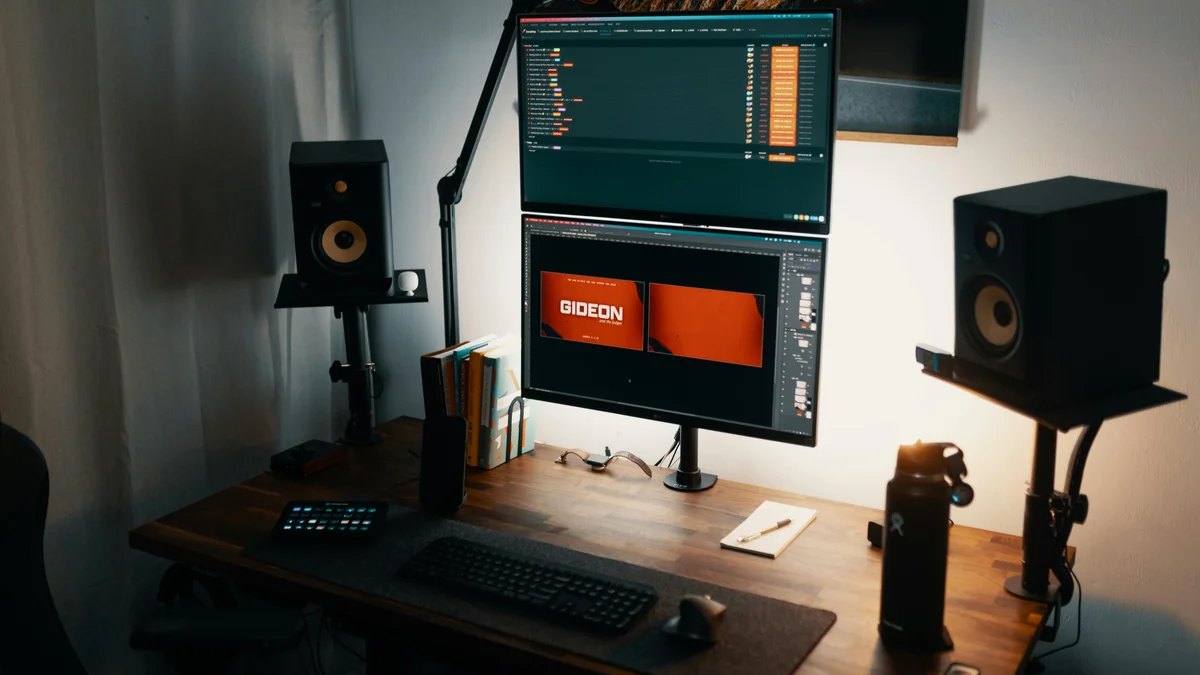 Espaço de trabalho moderno e bem iluminado, com dois monitores, colunas de som e uma mesa organizada.