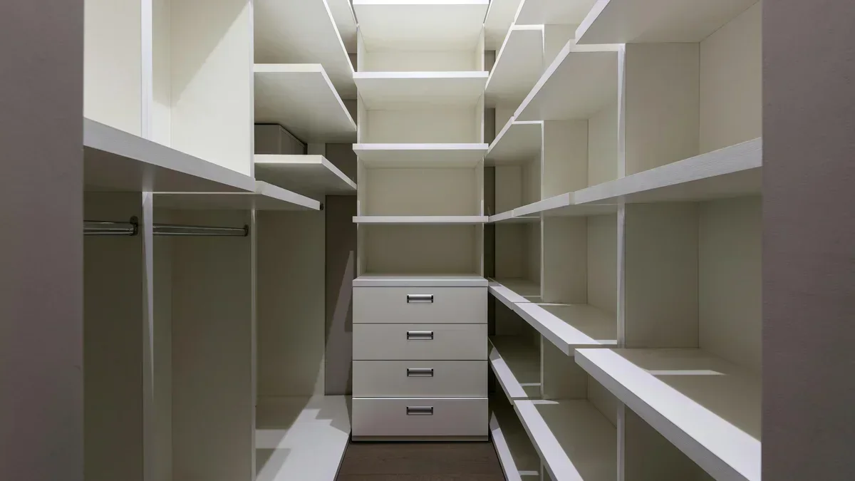 Prateleiras vazias e um armário alto com puxadores metálicos em um closet moderno e estreito, bem iluminado.