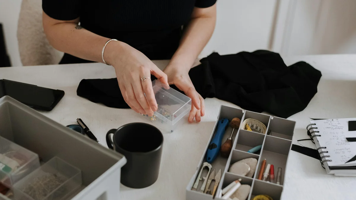 Mulher organizando materiais de costura em uma mesa de trabalho clara, com recipientes e caixa de alfinetes.
