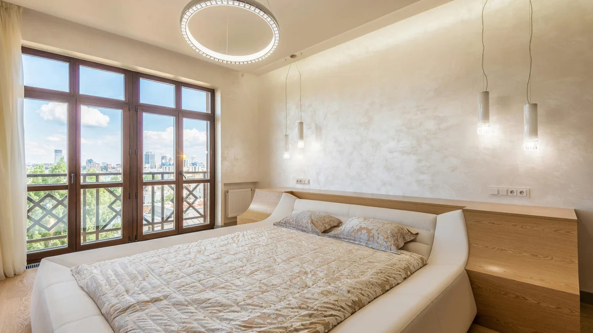 Interior de um quarto moderno com cama confortável, lustre e abajures, com janela e varanda, evidenciando um ambiente convidativo para o descanso.