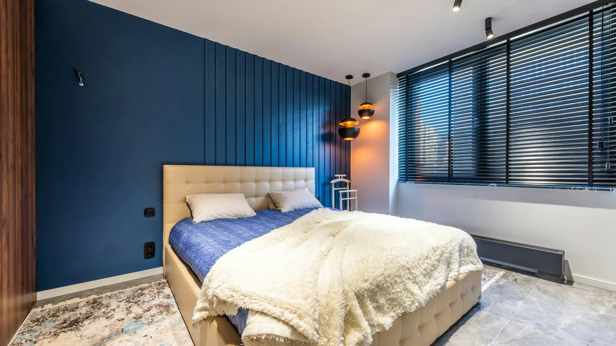 Cama com manta xadrez e almofadas, contra persianas e piso de ladrilho em um quarto contemporâneo, sugerindo conforto e estilo.