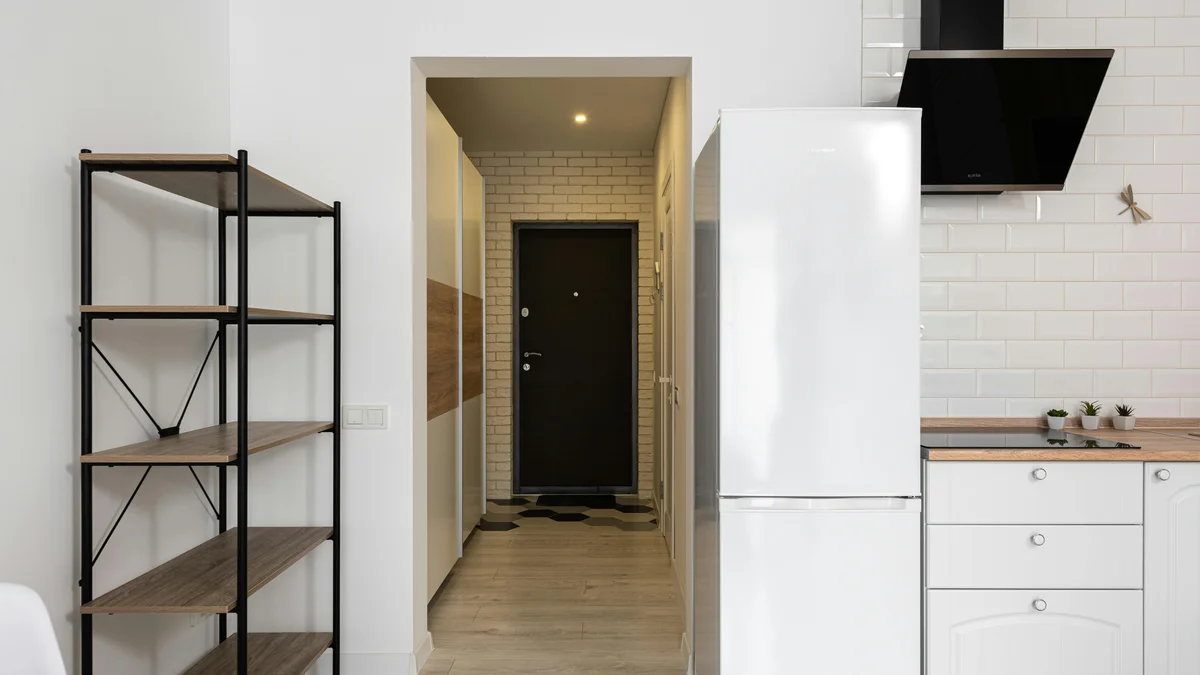 Cozinha moderna com geladeira encostada em prateleiras vazias, em uma casa com passagem estreita e porta durante o dia.
